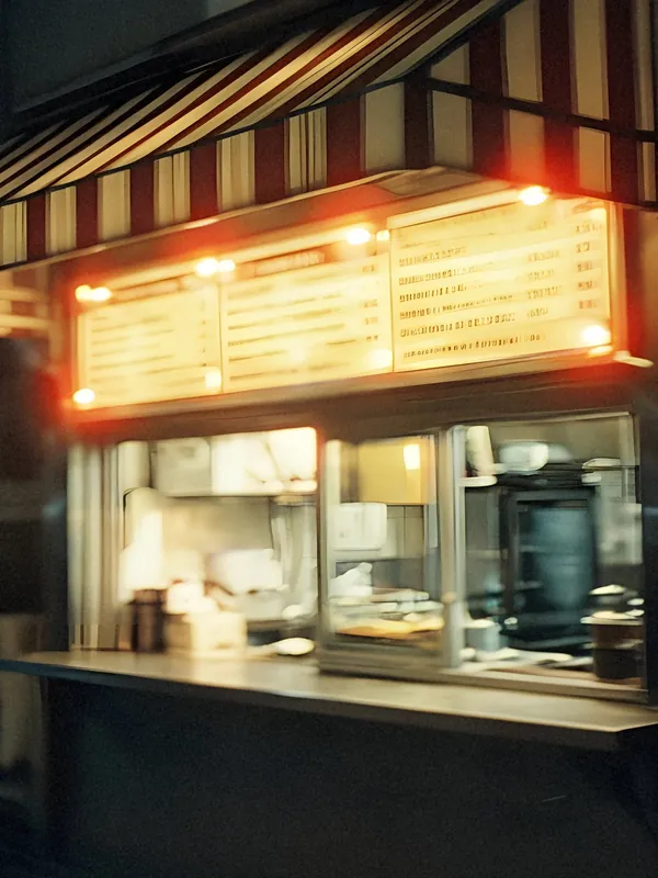 A taco stand window at night under a striped awning, the lit menu board above the counter